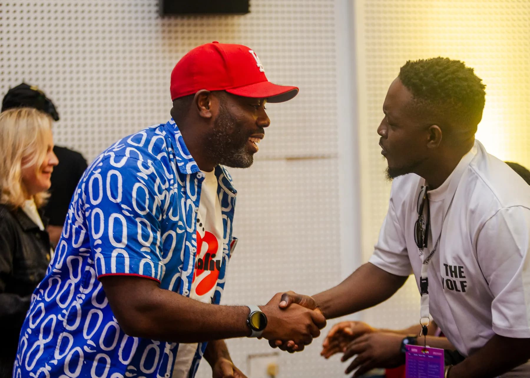 Two attendees in a warm handshake in the foyer, one wearing a red cap and a blue patterned shirt
