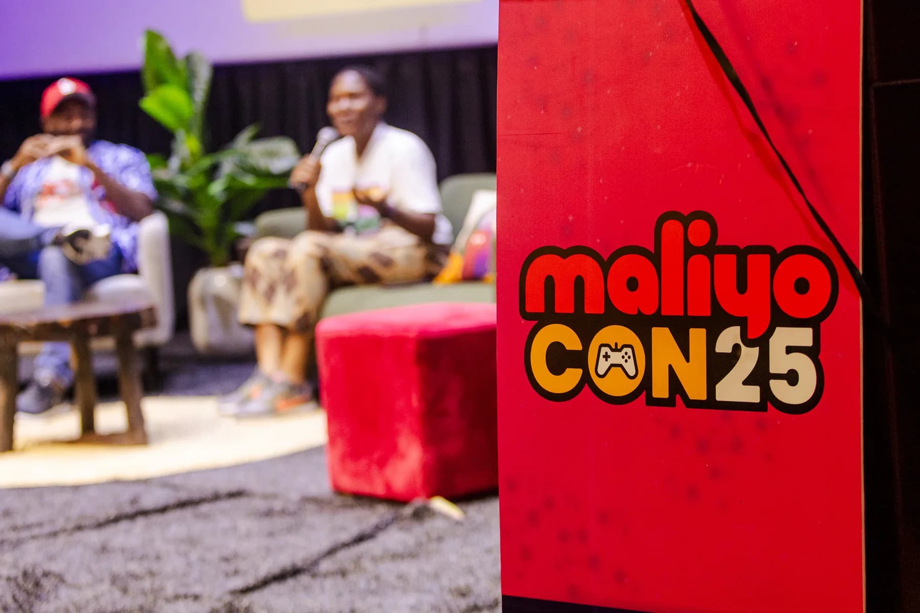 Fireside-style panel on stage with two speakers in armchairs, a red MaliyoCON25 banner in the foreground