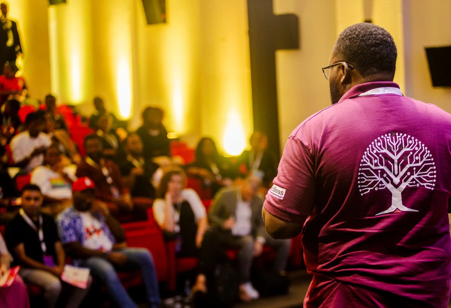 Audience seated in red chairs as a man in a maroon shirt with a tree logo stands in the foreground