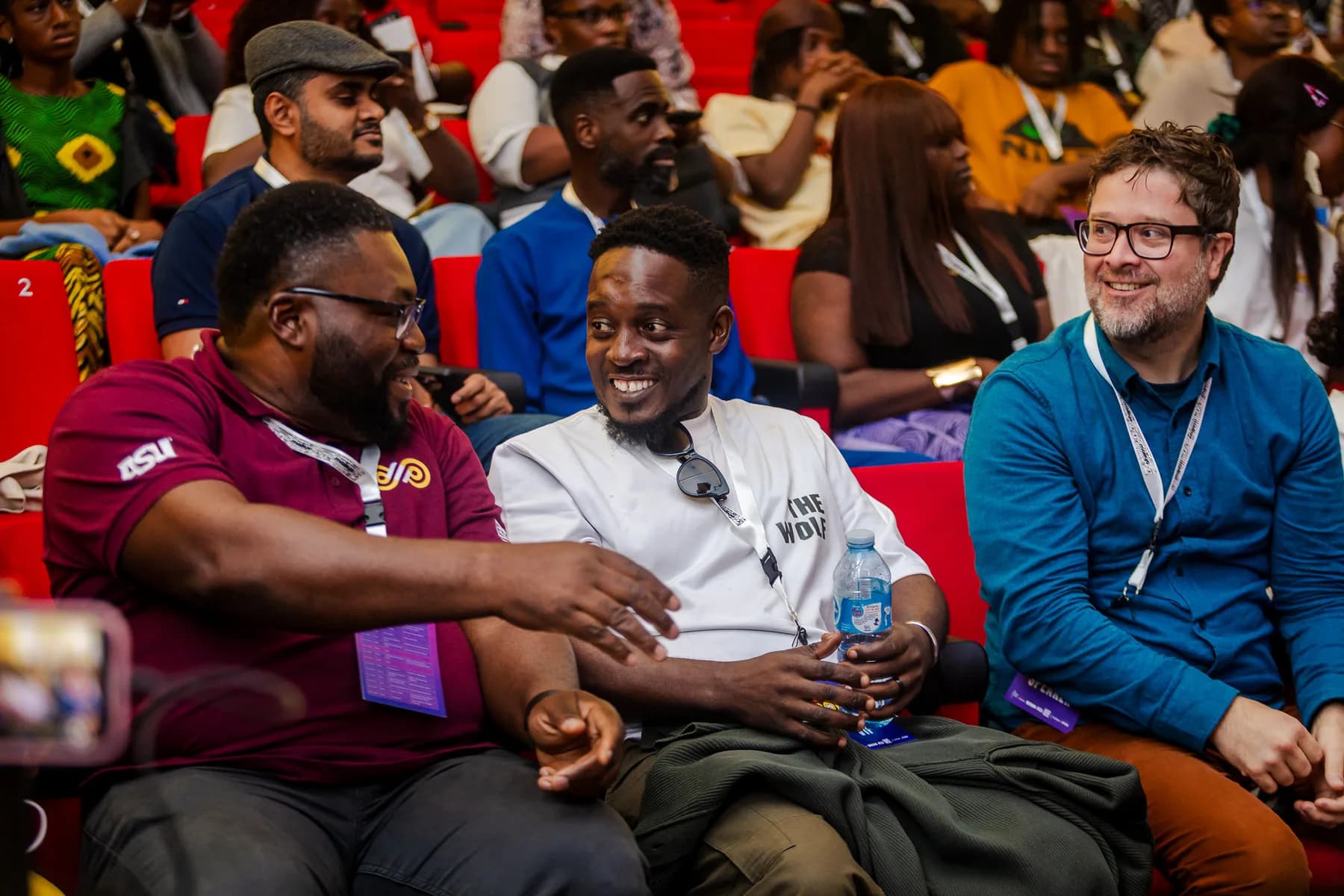 Three attendees sitting together in the audience, laughing and chatting, with conference lanyards
