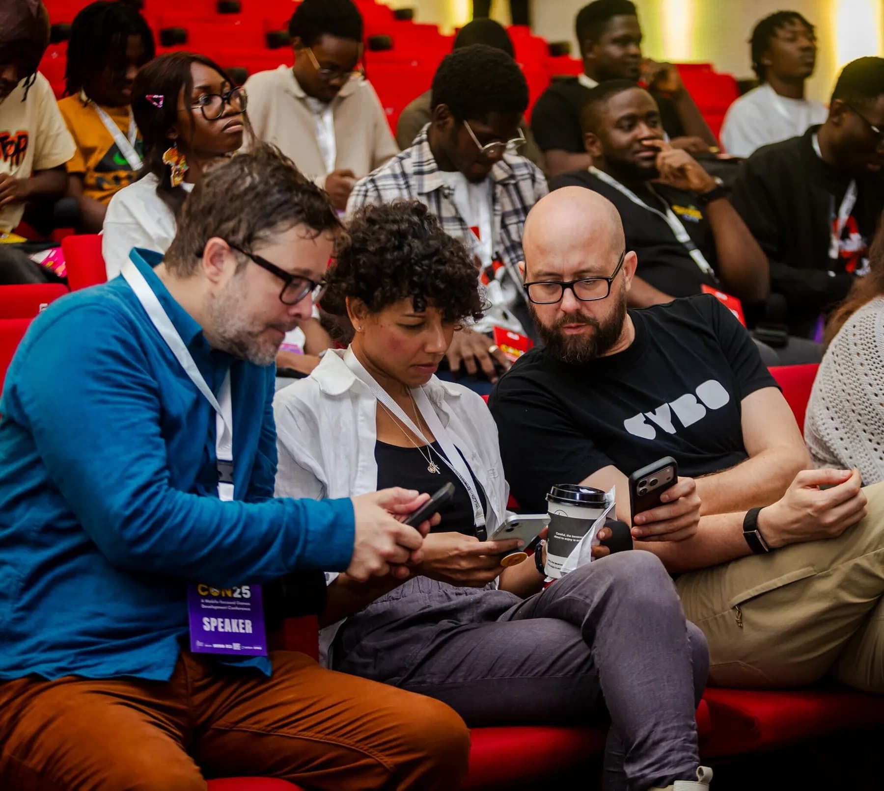 Three speakers in the audience looking at a phone together, one in a SYBO shirt, wearing speaker badges