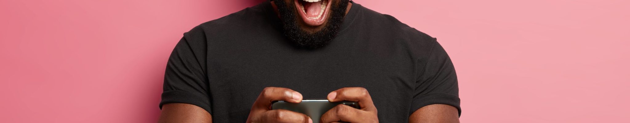 Excited bearded man holds modern smartphone horizontally, plays online game, uses heeadset, stuck at gadget screen, dressed in black t shirt, has fun alone, isolated on pink wall, has free time